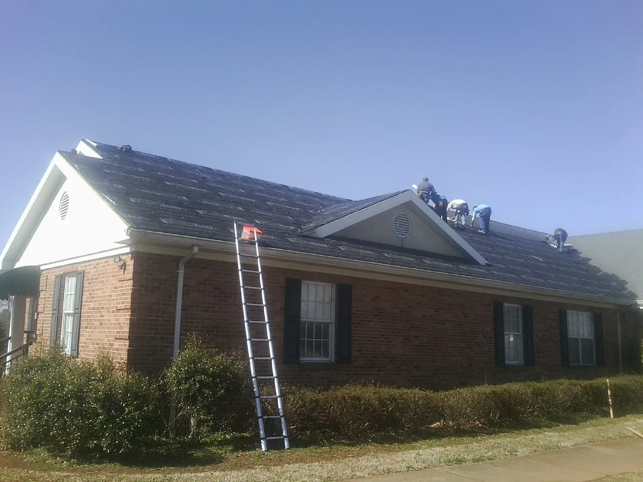 Crew tearing shingles off a brick ranch roof with a ladder set — Emergency Roof Repair work by our crew in Pinckney