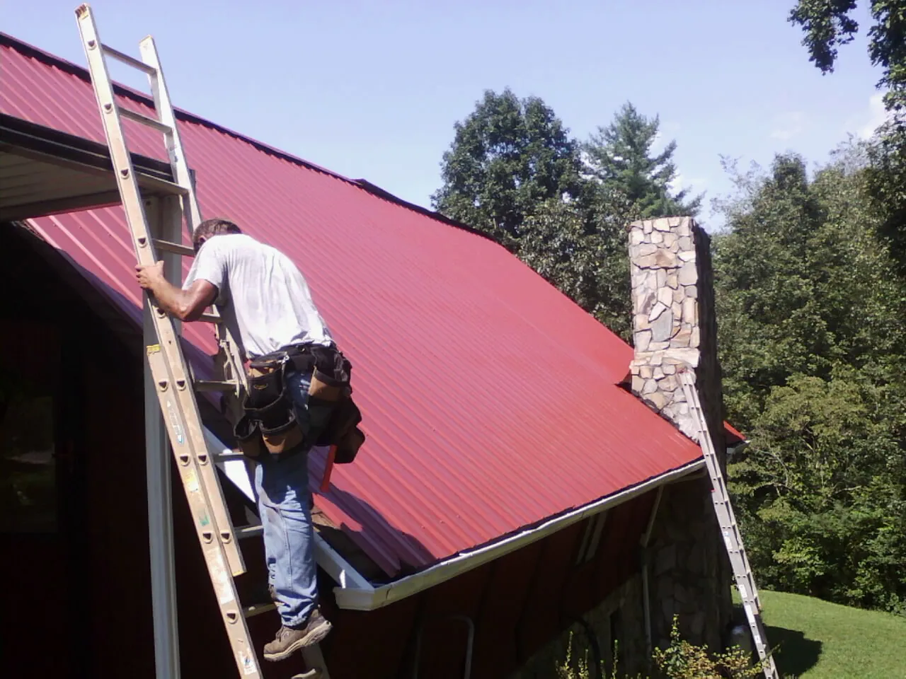 Roofer on a ladder beside a red metal roof and stone chimney — Soffit & Fascia Repair by Roofing Brothers in Pinckney